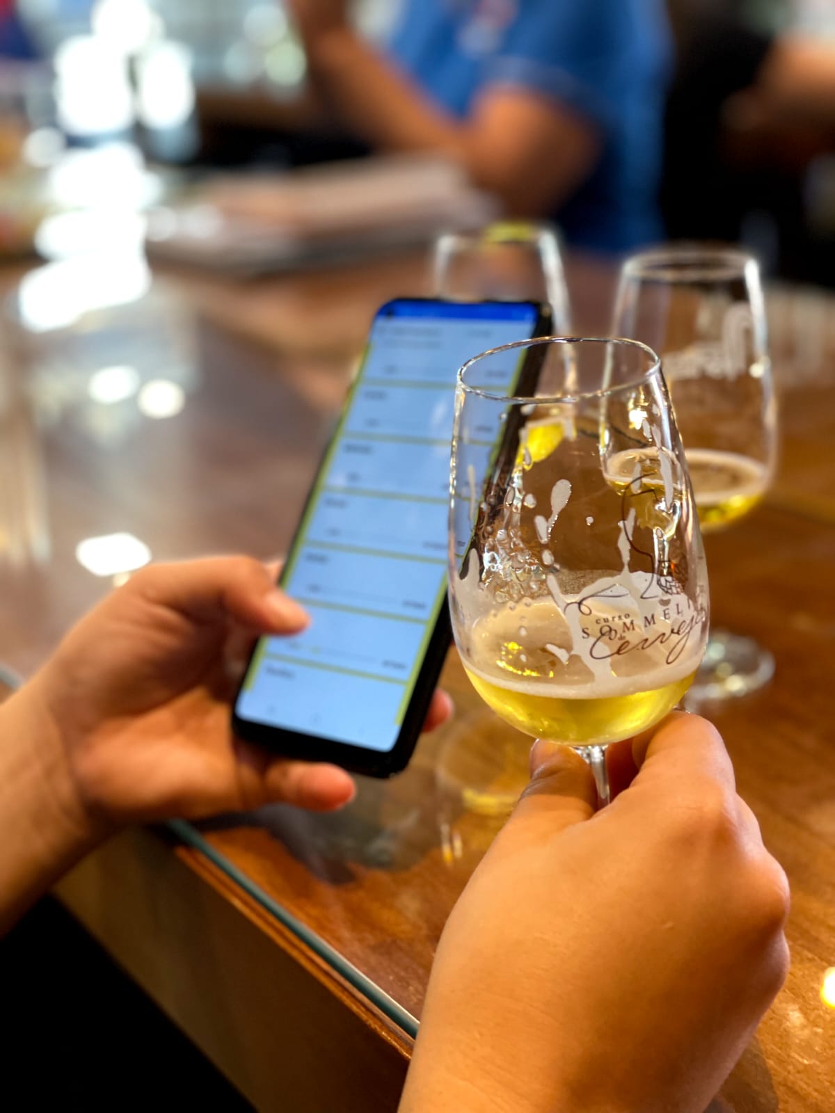 Imagem de uma pessoa bebendo uma taça de cerveja e com um celular na outra mão.
